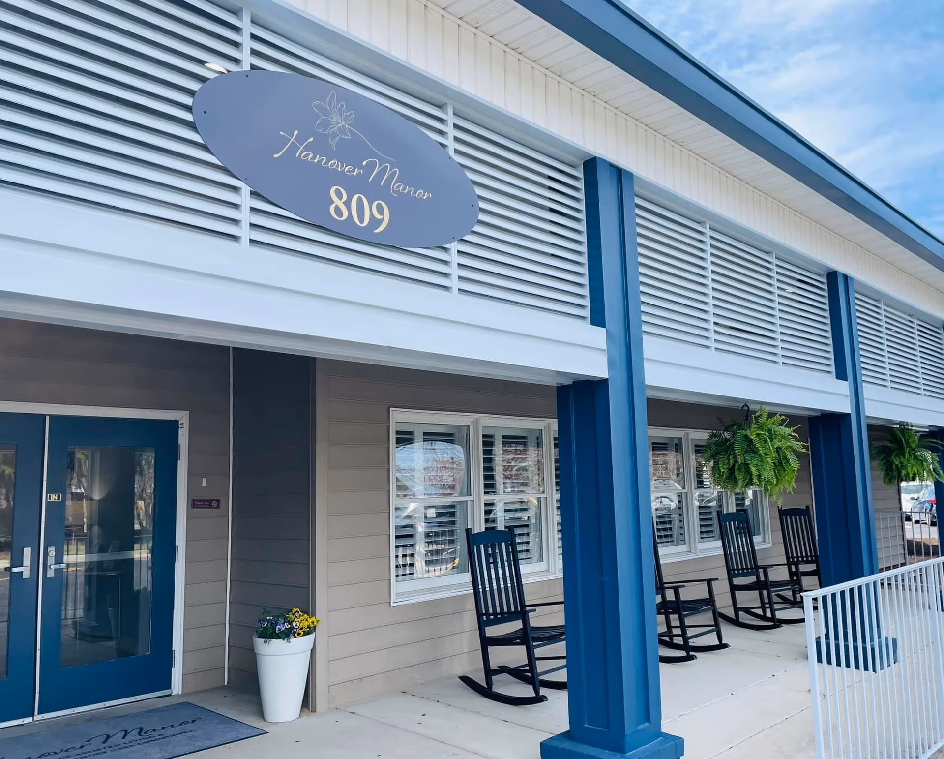 Exterior view of Hanover Manor Assisted Living building entrance with blue double doors, a sign displaying 'Hanover Manor 809', several black rocking chairs on the porch, hanging green plants, and a white flower pot with colorful flowers near the door.