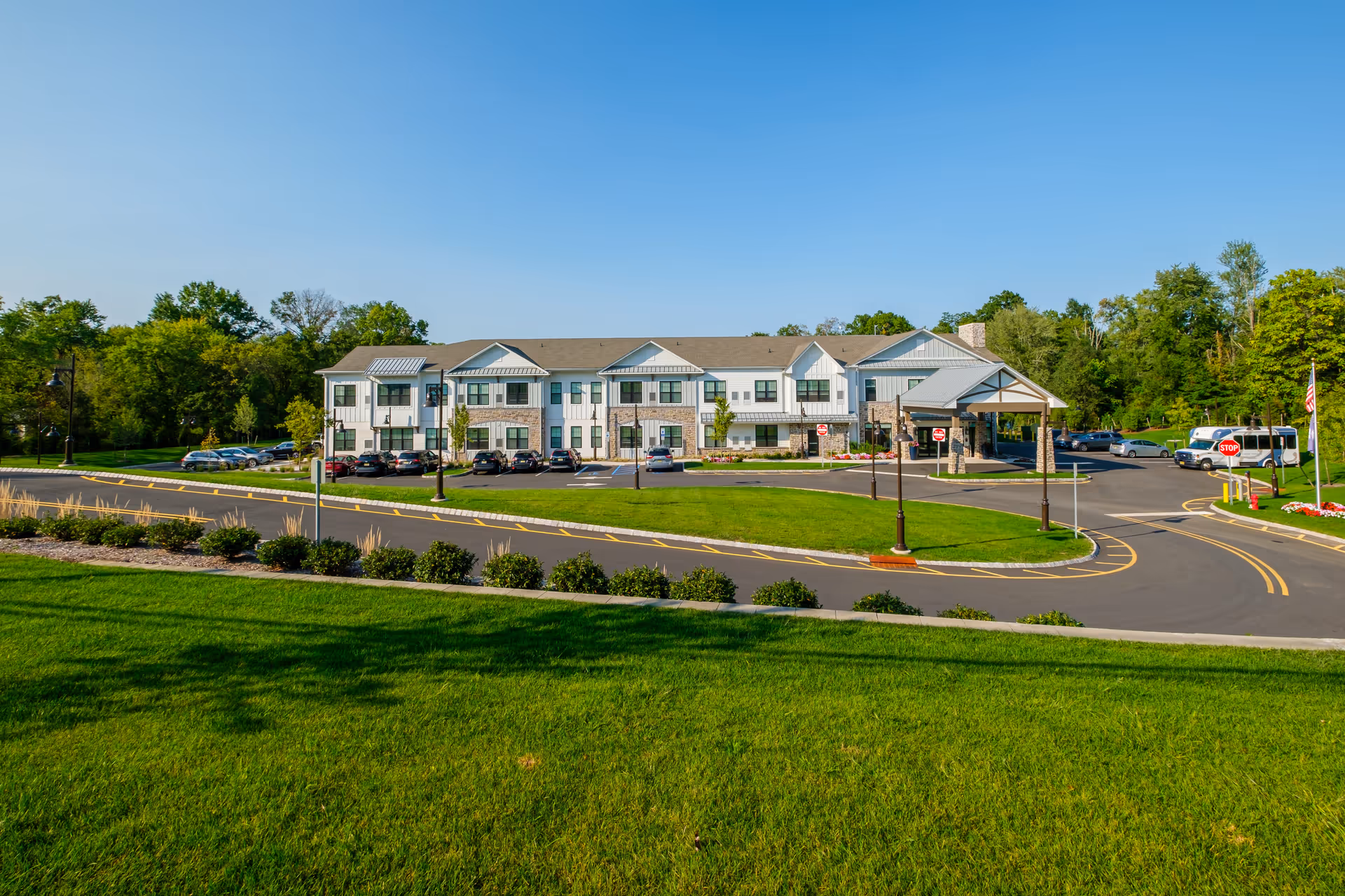 Exterior view of Arbor Terrace Basking Ridge, a two-story senior living facility with a large parking area, green lawns, and trees surrounding the building under a clear blue sky.