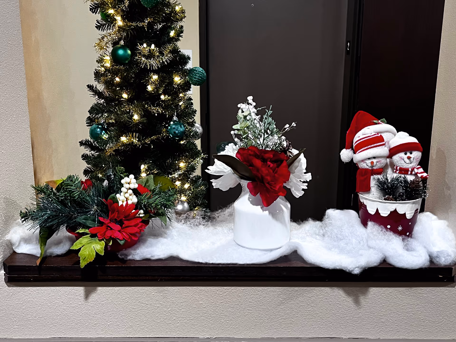 Indoor holiday arrangement with a Christmas tree, flowers, a white vase, and two snowmen.