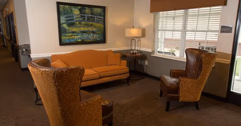 Small sitting area with an orange sofa, two patterned armchairs, a side table with a lamp, and a framed painting by a window.
