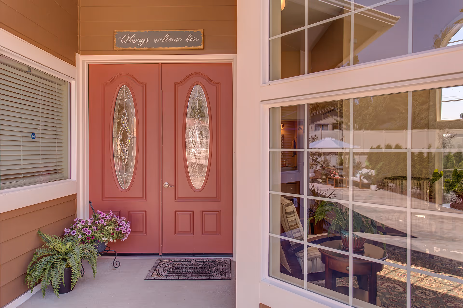 Double pink front doors with oval glass panels, Always welcome here sign, potted plants outside.