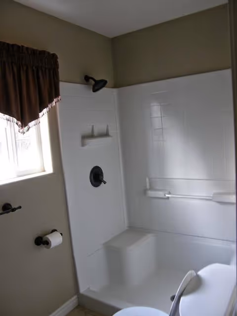 White tiled bathroom with shower, grab bars, toilet, and a small window.
