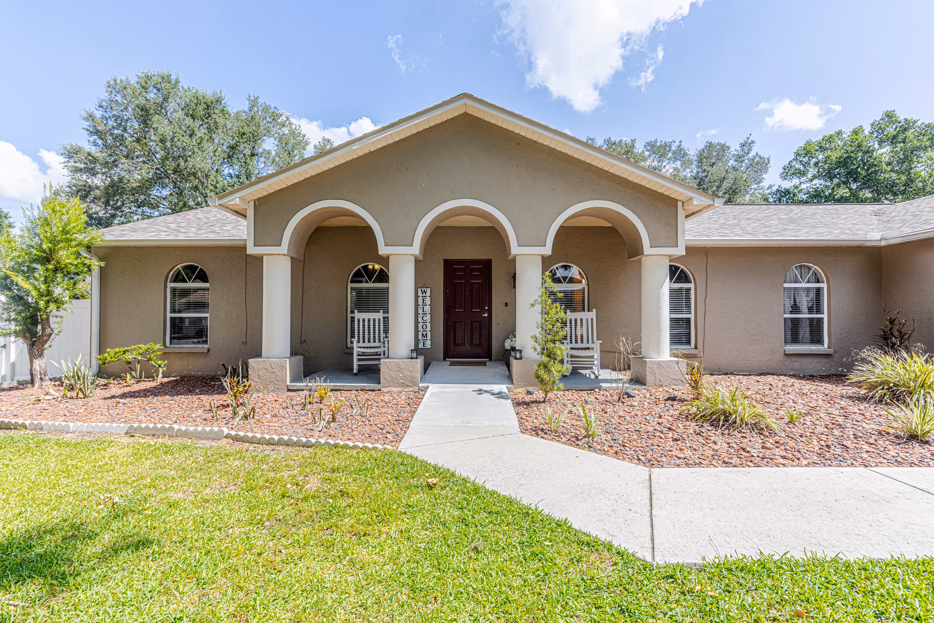A single-story senior living facility front entrance with arches, rocking chairs, and a landscaped walkway.