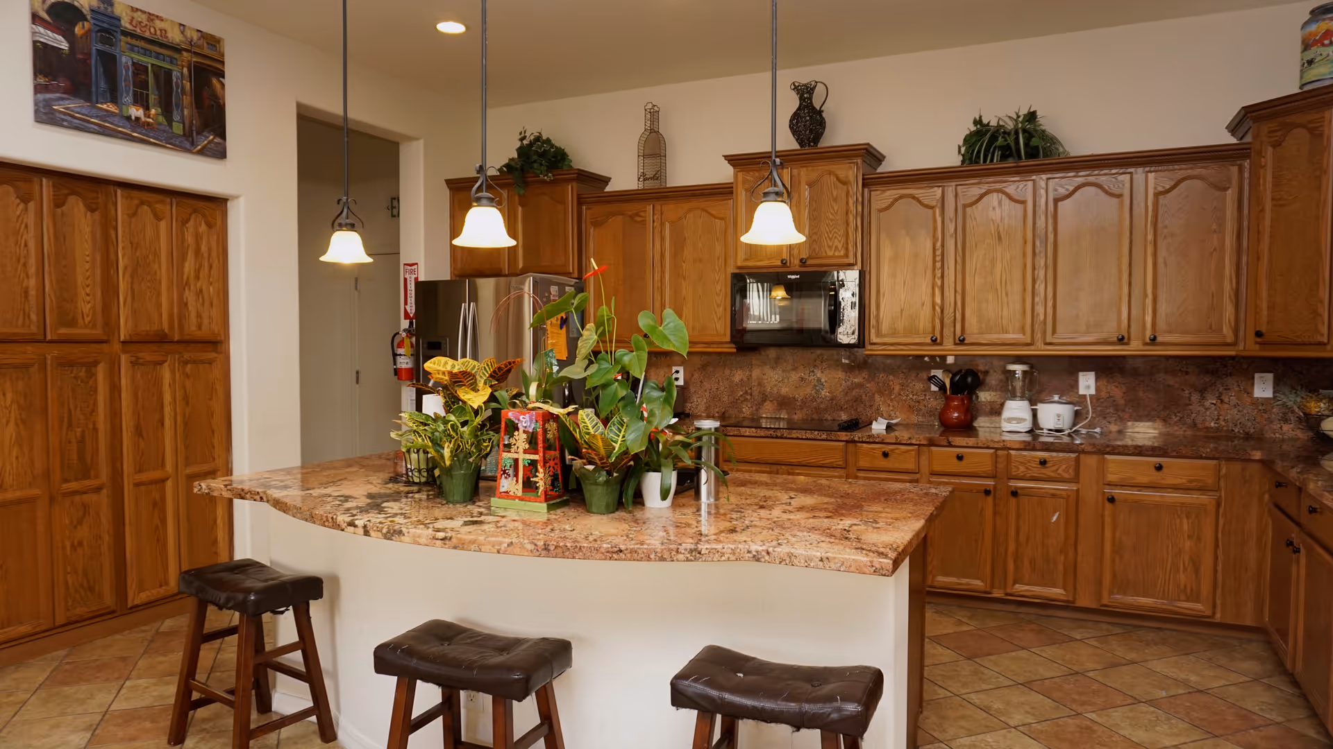 A spacious kitchen with wooden cabinets and a large granite countertop island. Three pendant lights hang above the island, which has three cushioned stools. Several potted plants and decorative items are placed on the island. The kitchen features a stainless steel refrigerator, a microwave, and various small appliances on the countertops. The floor is tiled, and there is a painting on the wall above a set of wooden doors.