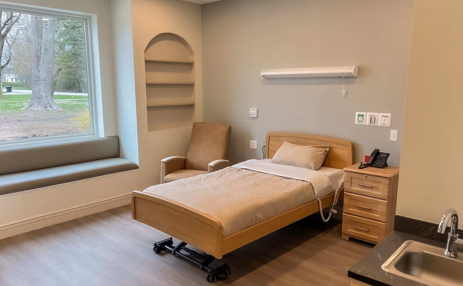 A single hospital-style private bedroom with a bed, nightstand, chair, and window view.