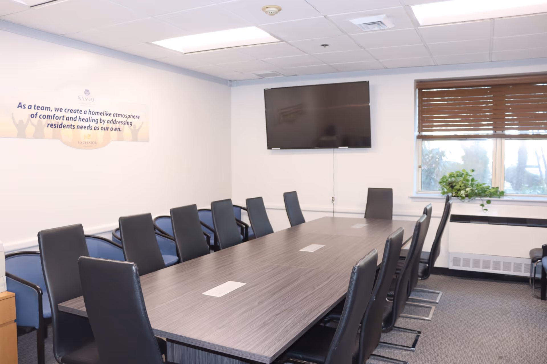 A long conference table with black chairs in a bright meeting room, TV mounted on wall.