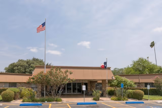 Single-story building entrance with American and Texas flags, parking spaces, and landscaped shrubs under a clear sky.