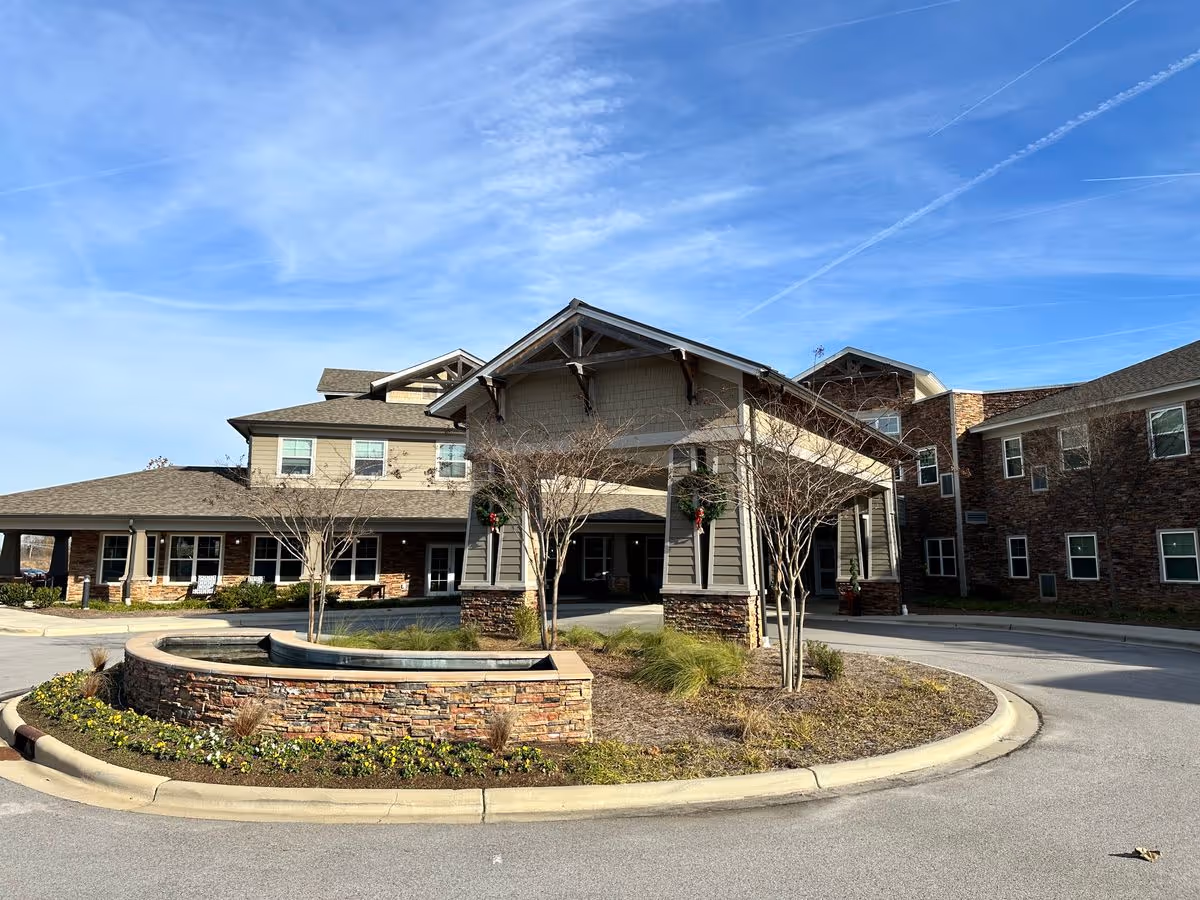 Front entrance of a multi-story senior living building with a covered porte-cochere, circular driveway, and a stone planter/fountain under a blue sky.