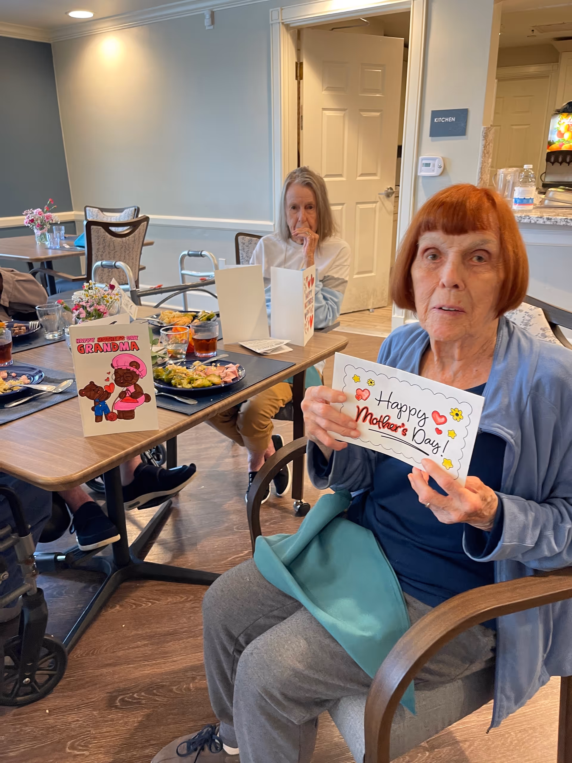 Two elderly women sit at a Senior Helpers dining table, holding a Mother's Day card.