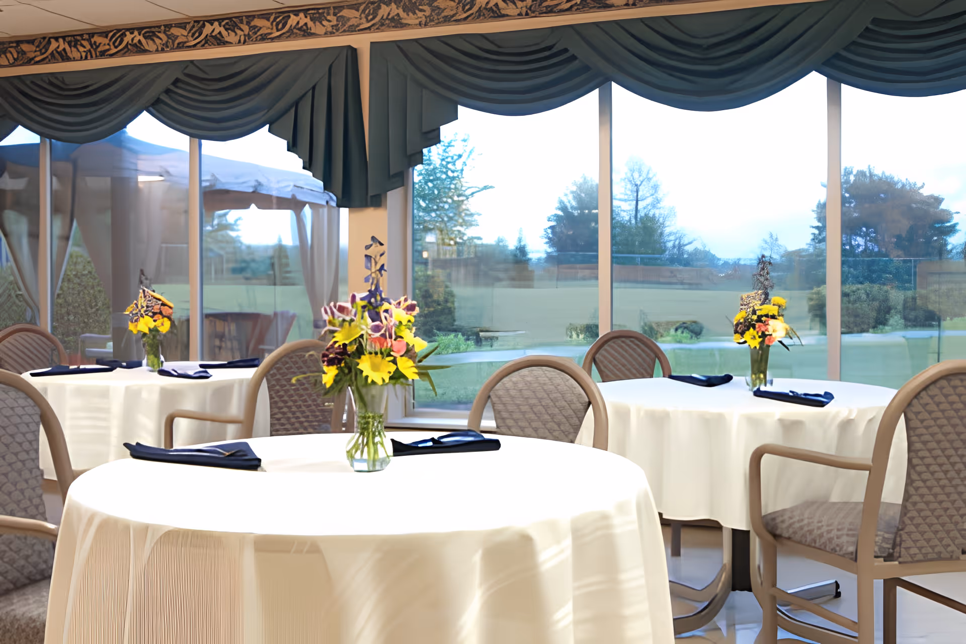 Dining room with round tables covered in white tablecloths, floral centerpieces, chairs, and large windows overlooking a lawn and gazebo.