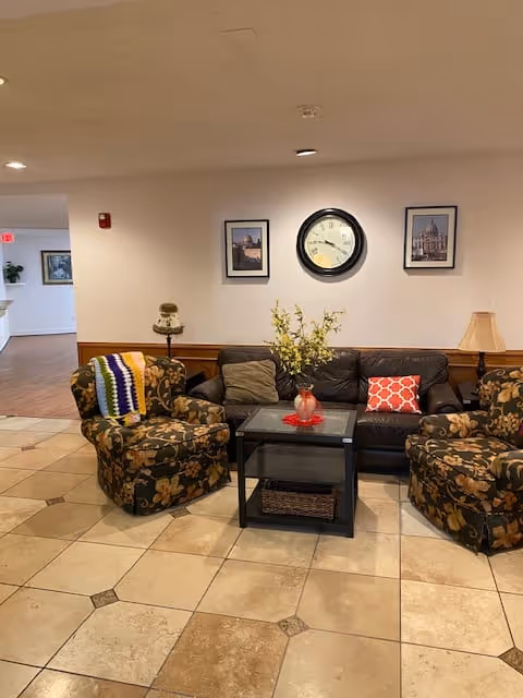 A cozy seating area with a dark brown leather sofa and two floral-patterned armchairs arranged around a black coffee table. The coffee table has a red vase with yellow flowers. On the wall behind the sofa, there is a round clock flanked by two framed pictures. The floor is tiled with large beige tiles, and there is a lamp on a side table to the right of the sofa.
