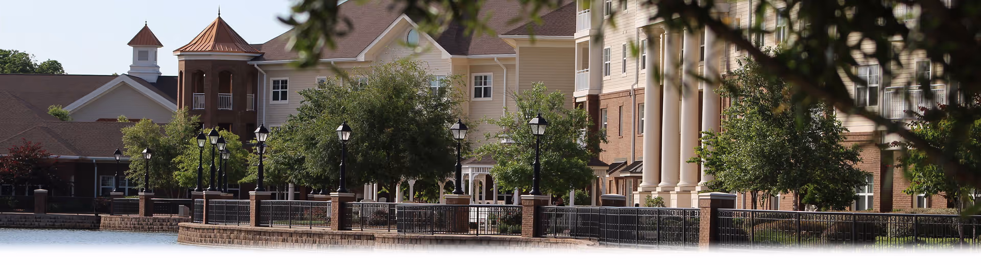Exterior view of a senior living facility named Live Oak, showing a waterfront with a stone retaining wall, black metal fencing, street lamps, trees, and a large building with beige siding, brick accents, and multiple windows under a clear sky.
