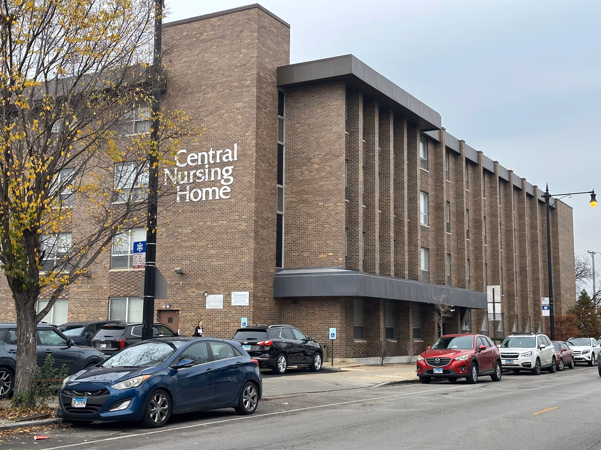 Exterior view of a multi-story brick building with the sign 'Central Nursing Home' on the side. Several cars are parked along the street in front of the building, and a tree with yellowing leaves is visible on the left side. The sky is overcast.