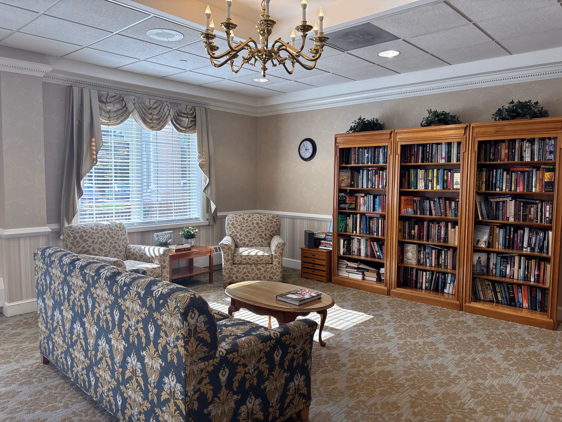 Cozy lounge area with patterned sofa, armchairs, bookshelf, and a window in soft lighting.