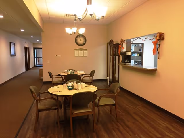 Interior view of a dining area in an assisted living facility with two round tables, each surrounded by four chairs. The tables are set with cups, plates, and condiments. A clock and a framed notice are mounted on the wall. There is a pass-through window to a kitchen area decorated with fall-themed garlands. The floor is wood, and the walls are painted light beige.