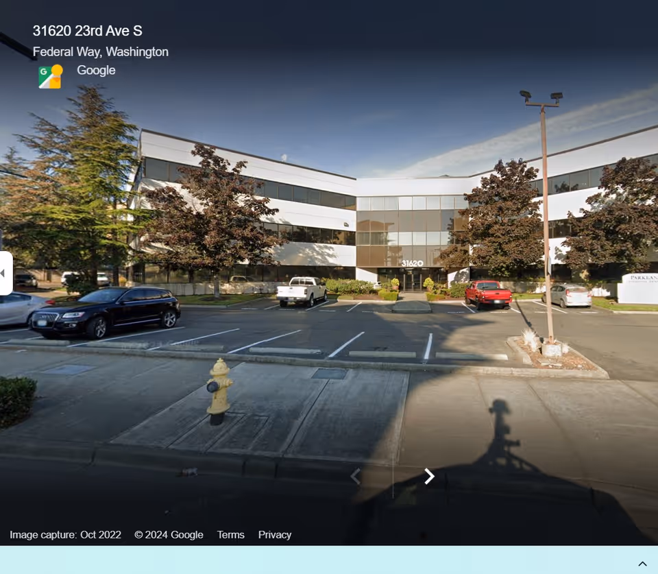 Front view of a modern office building with glass facade, parking lot, and trees.