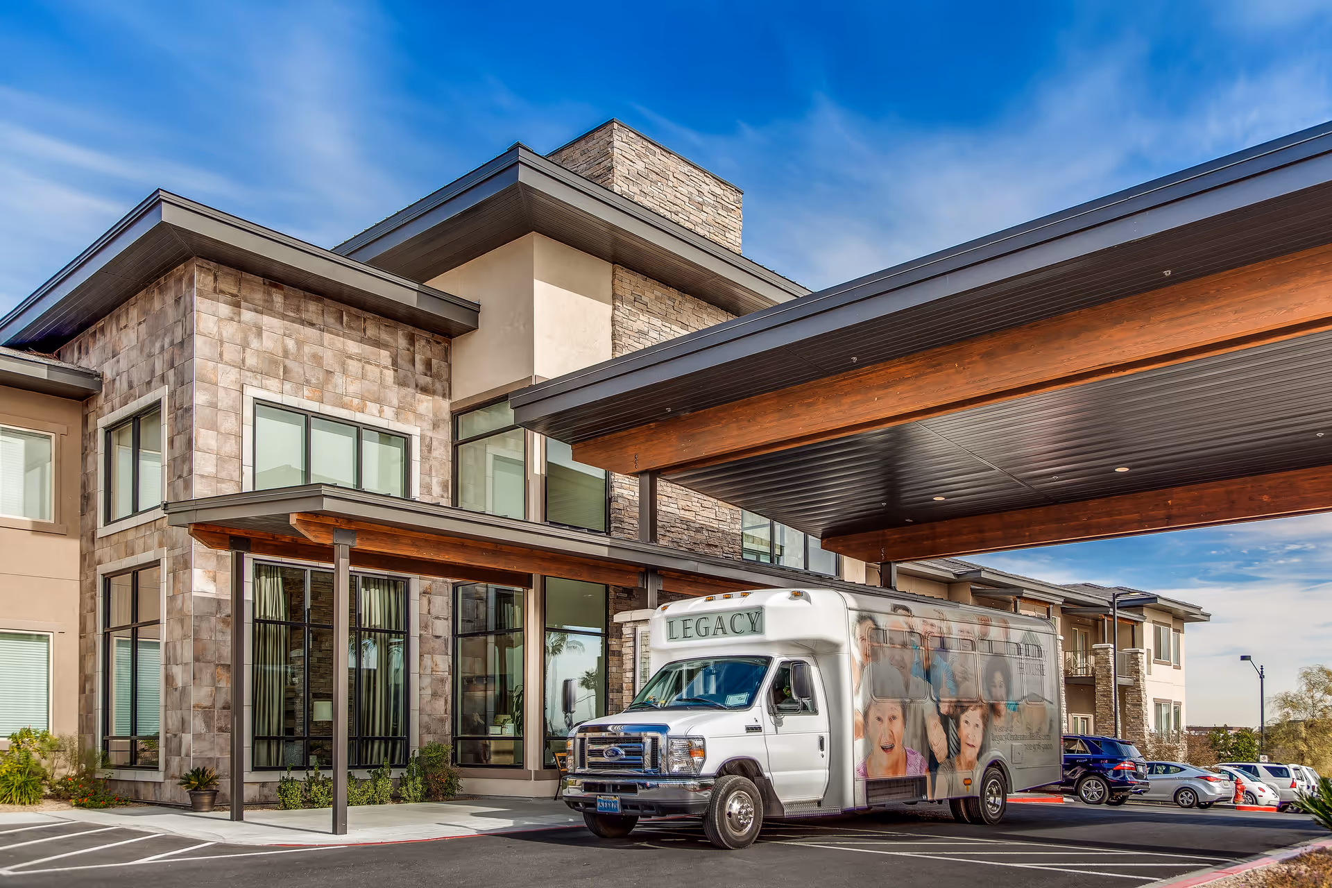 Exterior view of Legacy House of Centennial Hills building with a covered entrance and a white shuttle bus parked in front. The bus has images of elderly people on its side. Several cars are parked nearby under a blue sky with some clouds.