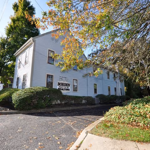 White, two-story building with trees and a sign reading BORDEN REALTORS in a parking lot.
