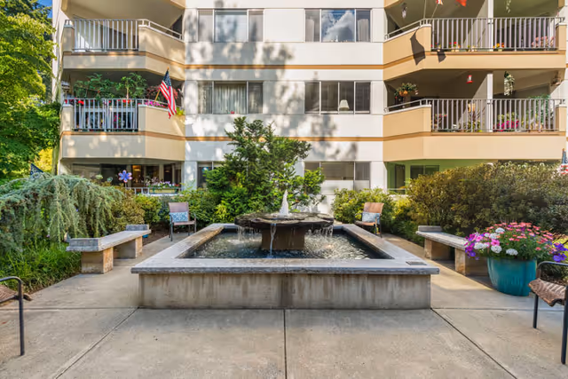 Outdoor courtyard area of a senior living facility with a central water fountain surrounded by benches, chairs, and lush greenery. The building facade with balconies and windows is visible in the background, along with an American flag hanging from one balcony.
