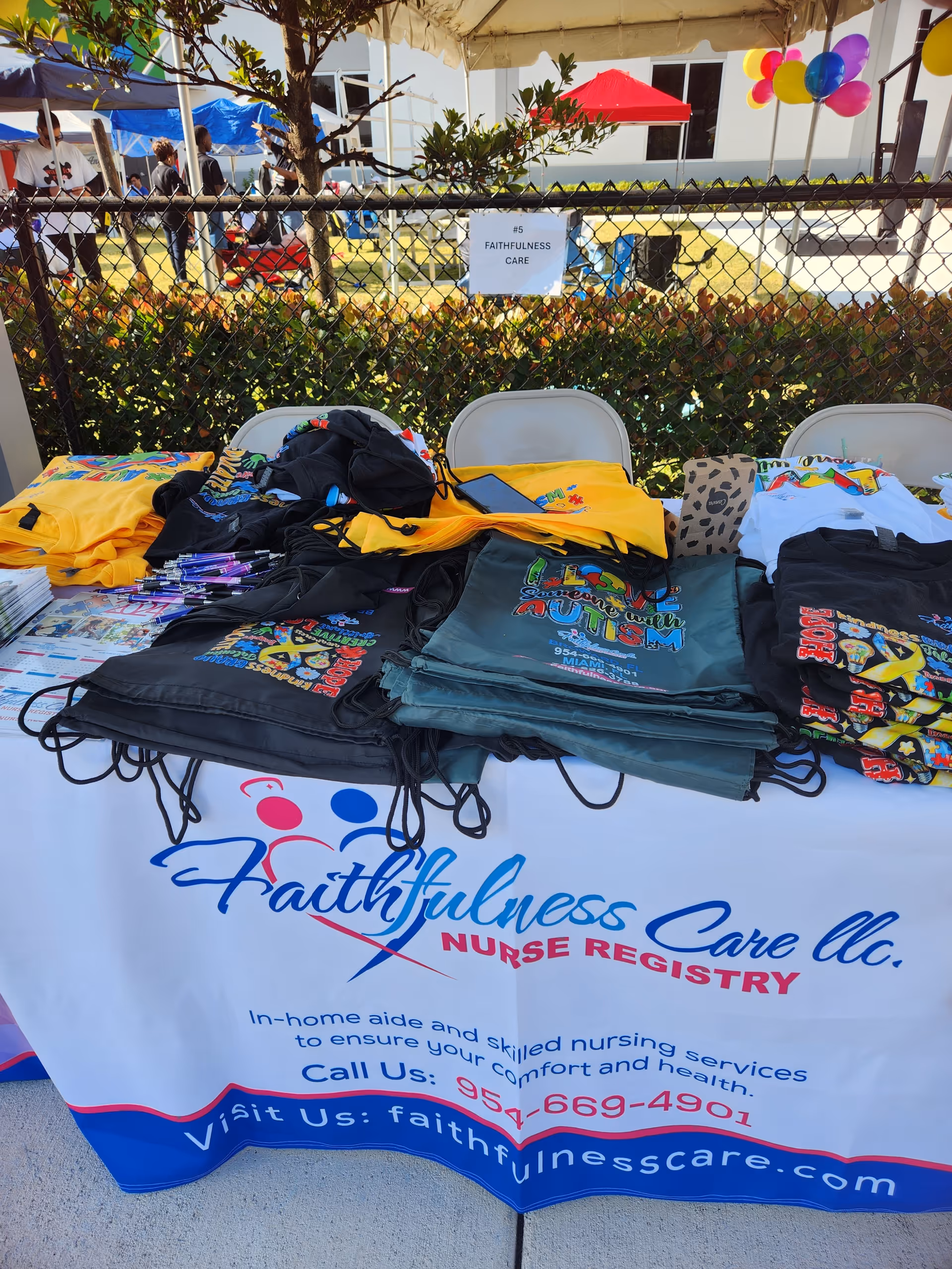 Outdoor table with Faithfulness Care LLC banners and stacked branded t-shirts under tents