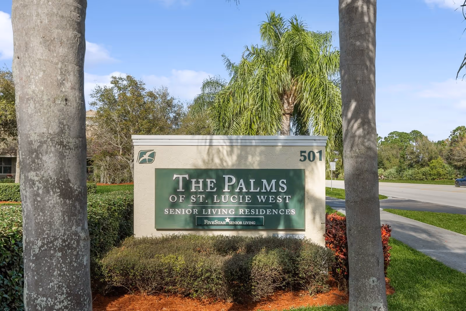Outdoor view of a large sign for The Palms of St. Lucie West Senior Living Residences, surrounded by landscaping with bushes and palm trees, located near a road under a partly cloudy sky.