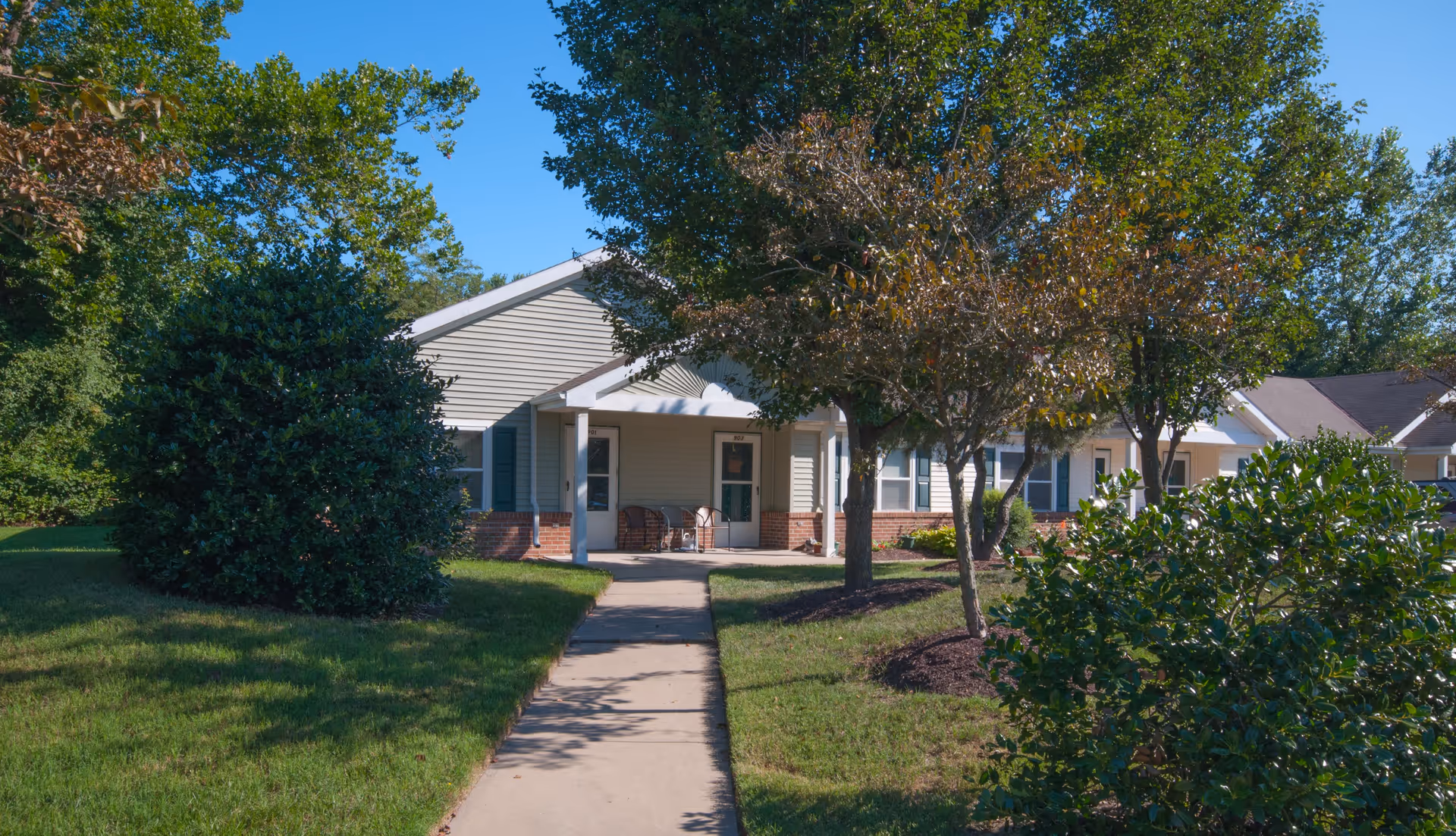 A single-story residential building with beige siding and brick accents, surrounded by green grass, bushes, and trees under a clear blue sky. A concrete pathway leads to a covered porch with chairs in front of two doors labeled 901 and 902.
