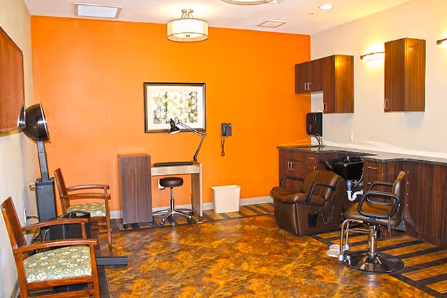 Interior view of a salon area in a senior living facility with two salon chairs, a small desk with a lamp and stool, wooden cabinets mounted on the wall, and two wooden chairs with floral cushions against an orange accent wall.