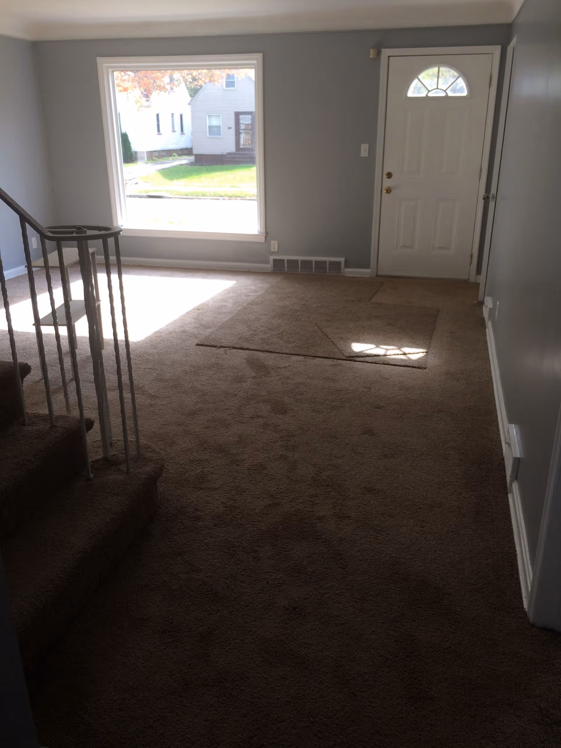 Sunlit empty interior room with window, staircase railing, and front door.