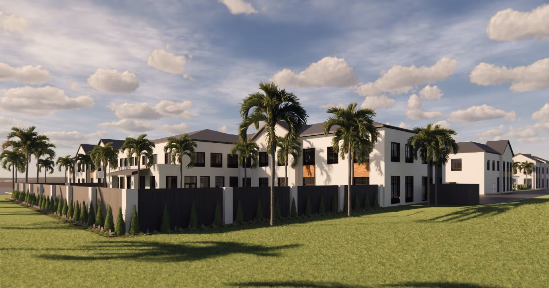 Exterior view of a modern two-story building with white walls and dark roofs, surrounded by palm trees and a black fence, under a partly cloudy sky.