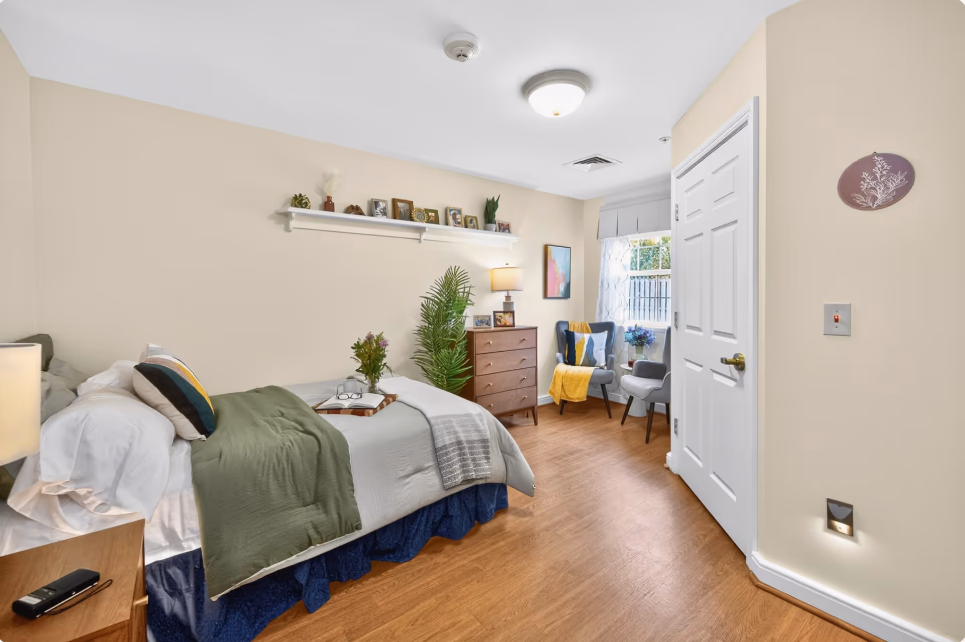 Cozy senior living bedroom featuring a bed, dresser, chairs, plants, and decorative wall shelf.