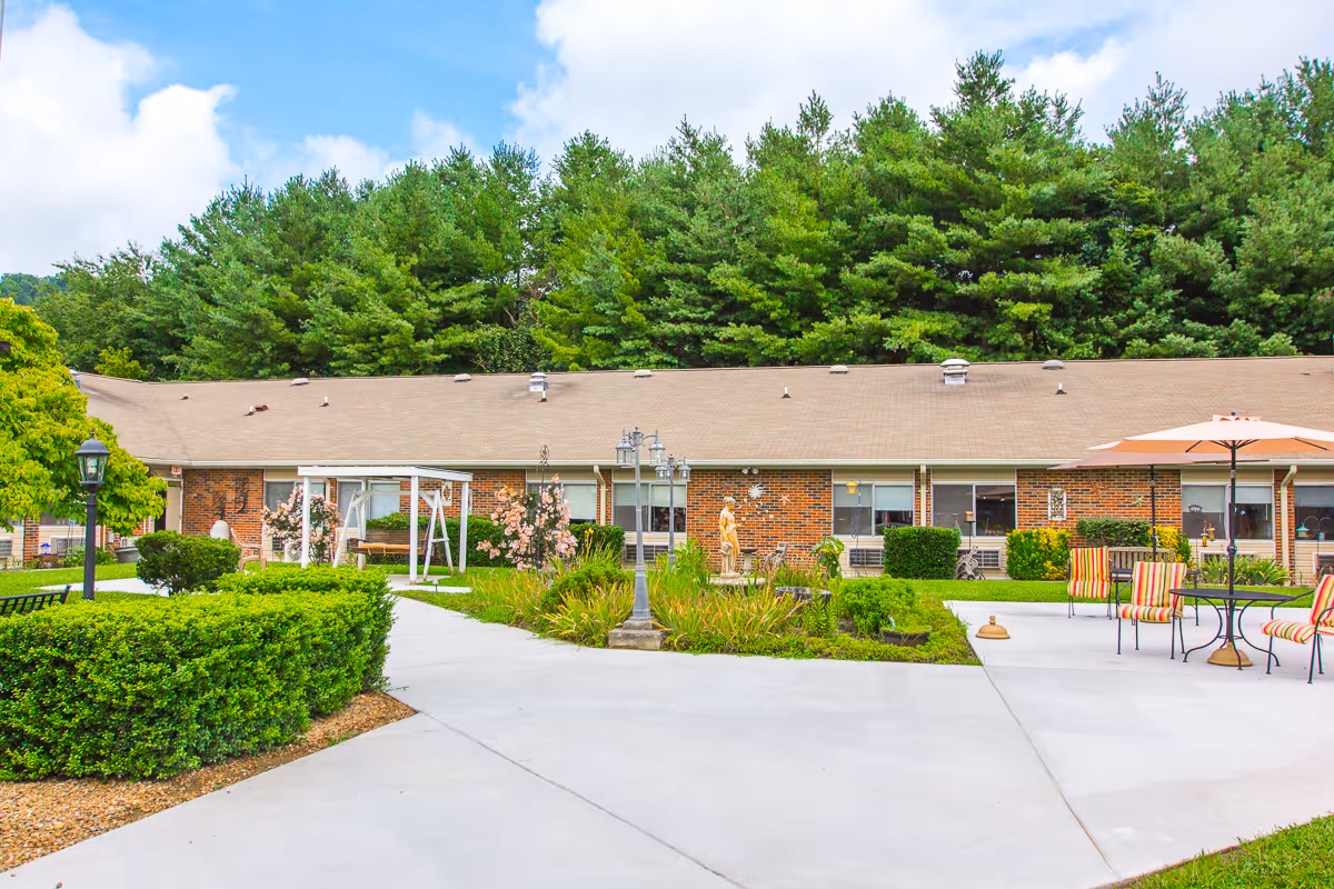Outdoor courtyard area of a senior living facility with a paved walkway, green bushes, a white swing, a statue, and patio tables with umbrellas and striped chairs. The building is a single-story brick structure with large windows, surrounded by tall green trees under a partly cloudy sky.