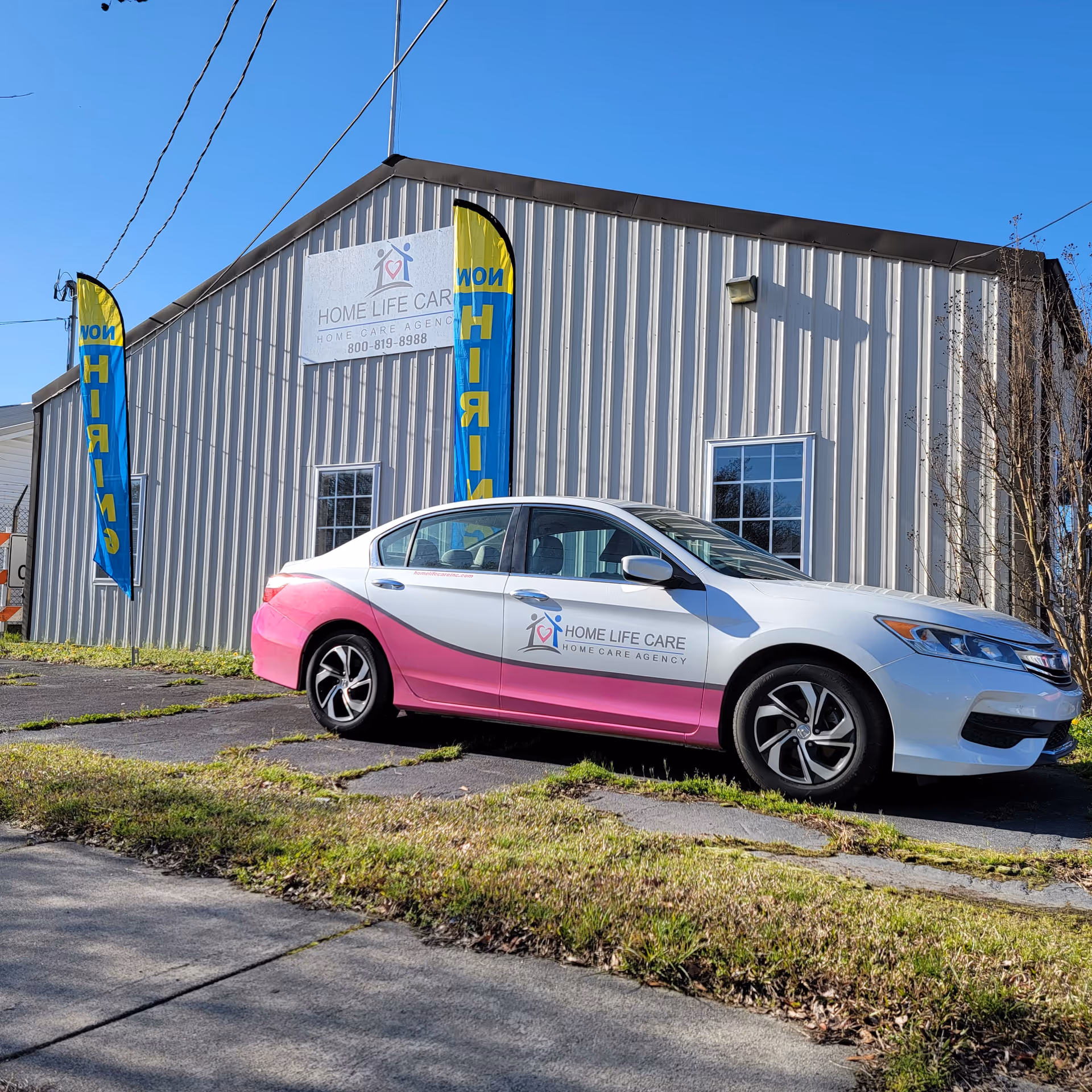 Exterior view of a metal-clad building with a branded Home Life Care car and flags.