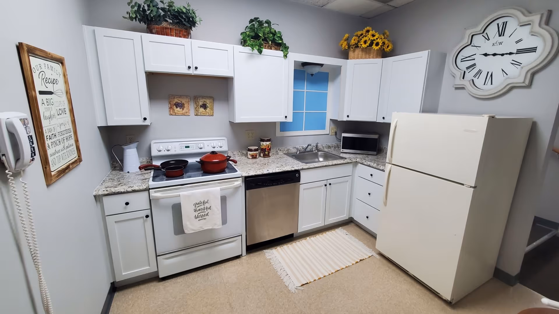 Small white kitchen with stove, refrigerator, sink, microwave, and decorative plants in a senior facility.