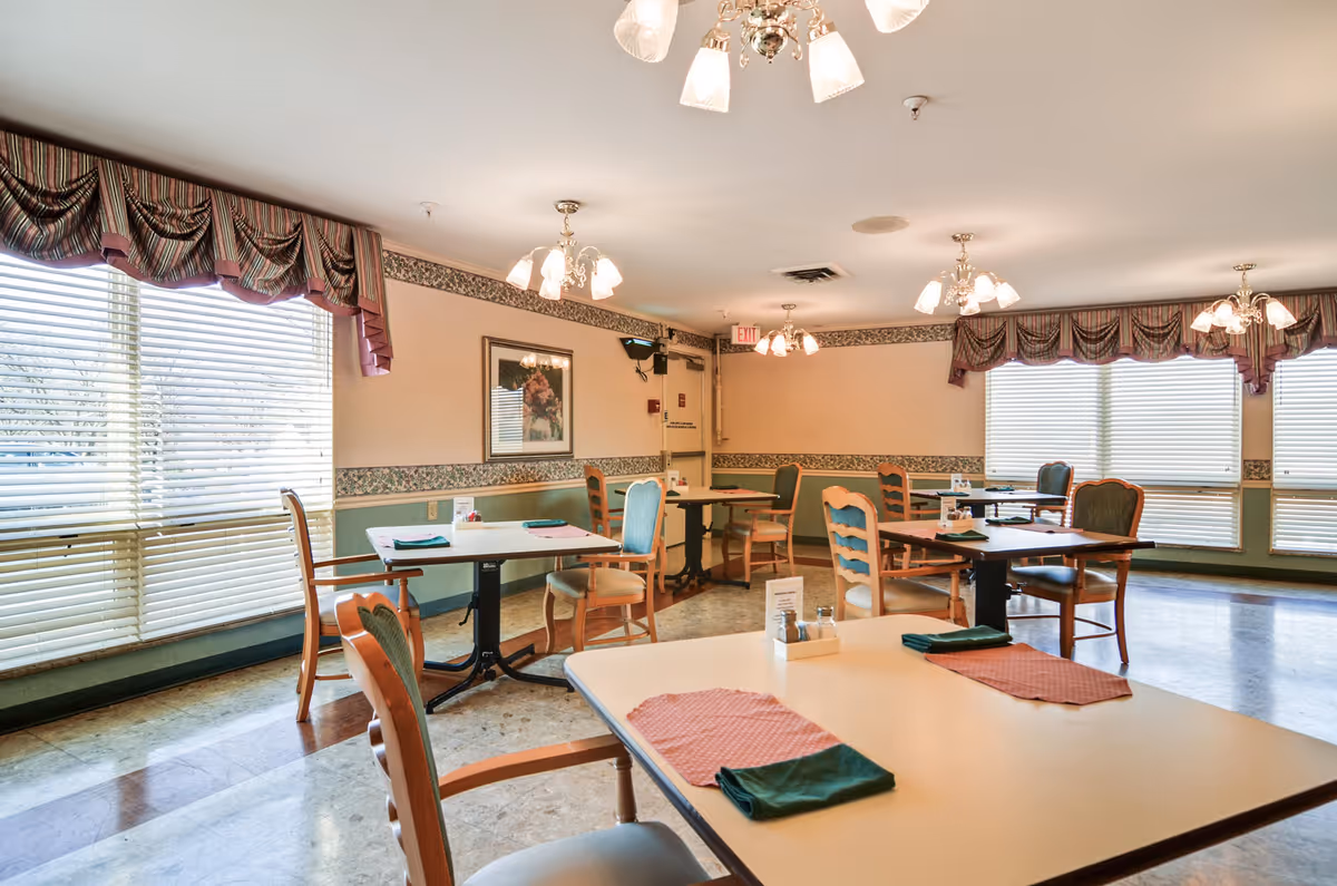 Interior of a senior living facility dining room with tables, chairs, and large windows.