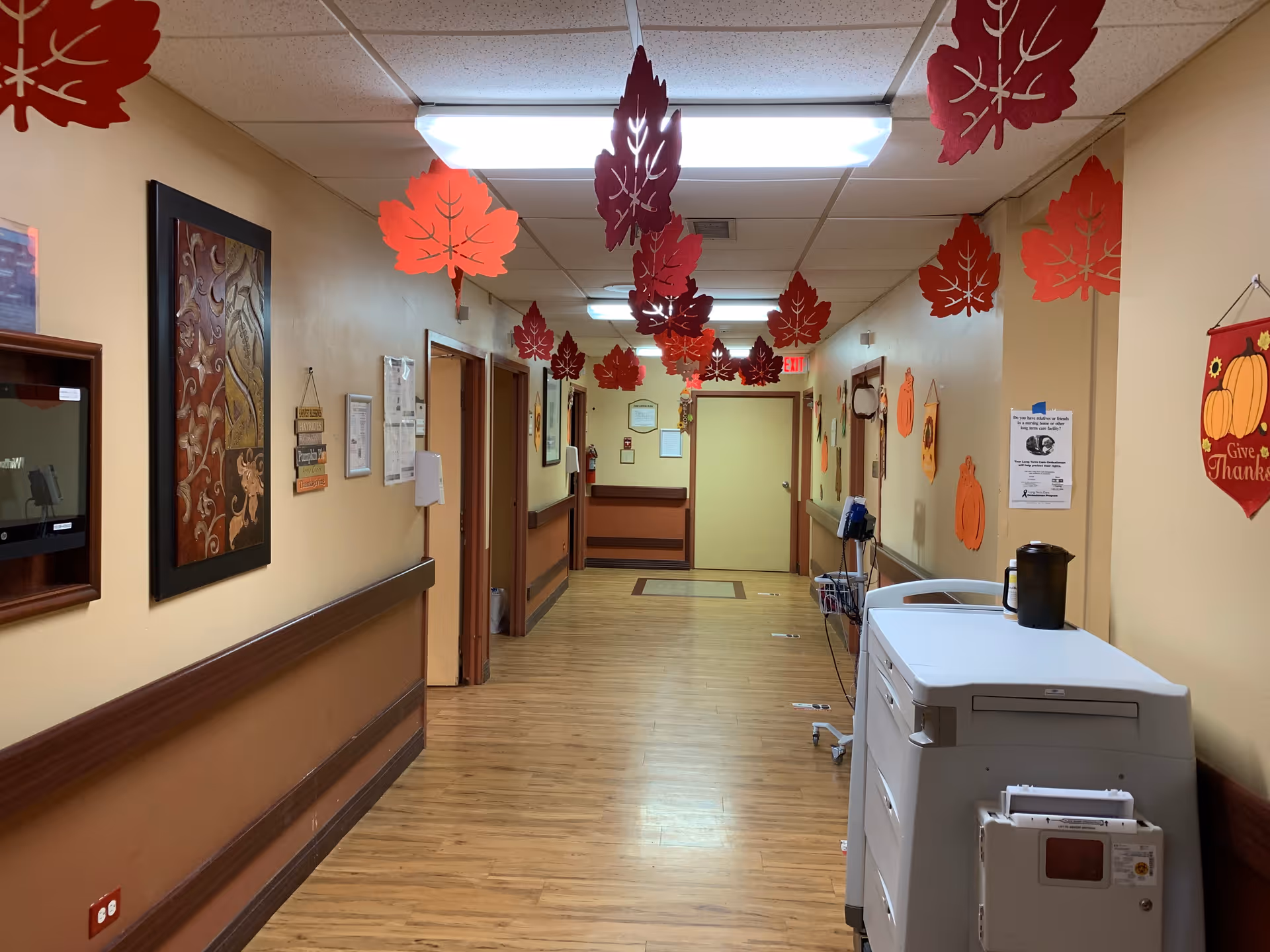 A hallway in a senior living facility decorated with hanging red and orange paper leaves from the ceiling. The walls are beige with brown handrails, and there are various decorations including autumn-themed wall hangings and framed artwork. A white medical cart with equipment is positioned on the right side of the hallway.