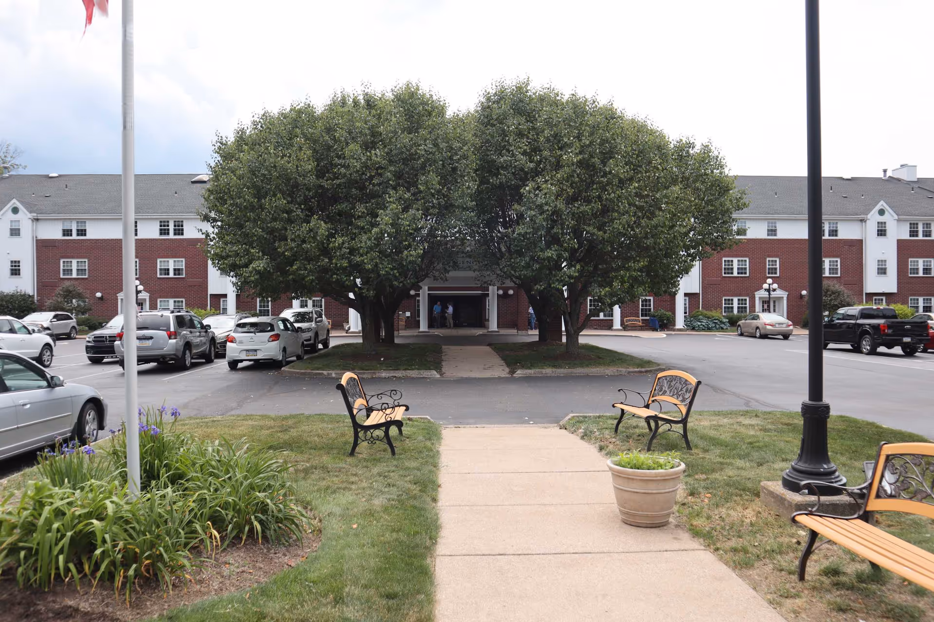 View of the entrance to Parkside Regency at South Shore senior living facility, showing a paved walkway flanked by benches and flower beds leading to the building entrance partially obscured by two large trees. Several parked cars are visible in the parking lot surrounding the building.