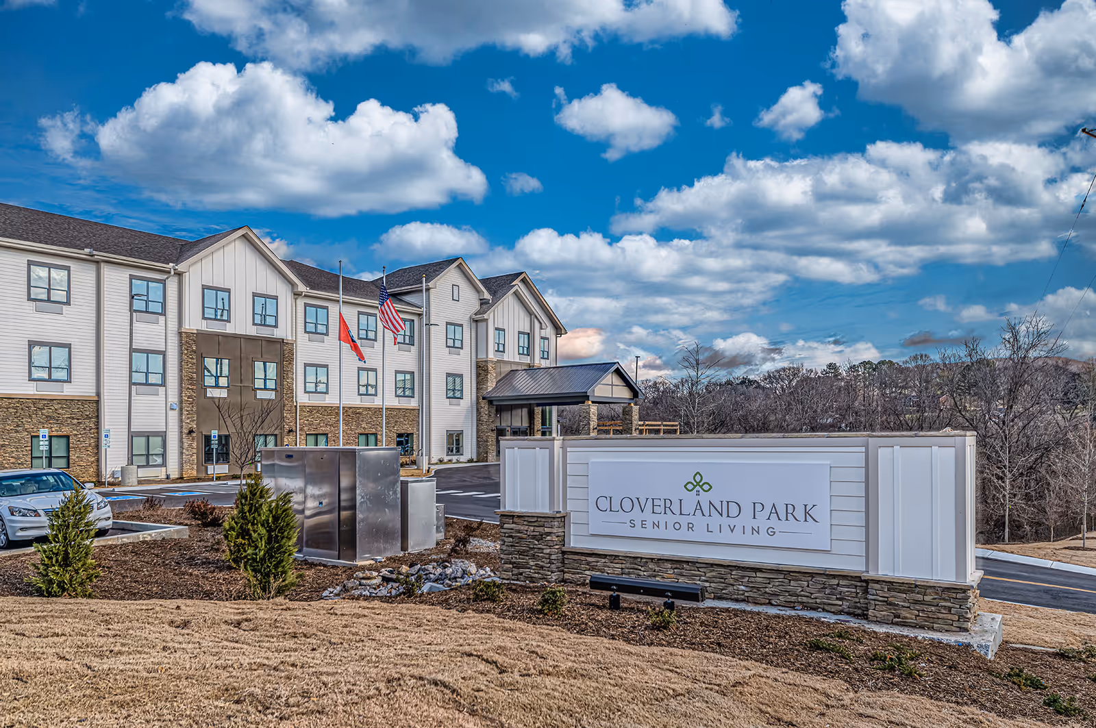 Front exterior of Cloverland Park Senior Living showing the building, entrance canopy, flagpoles, parking area, and a large sign on a landscaped lawn.
