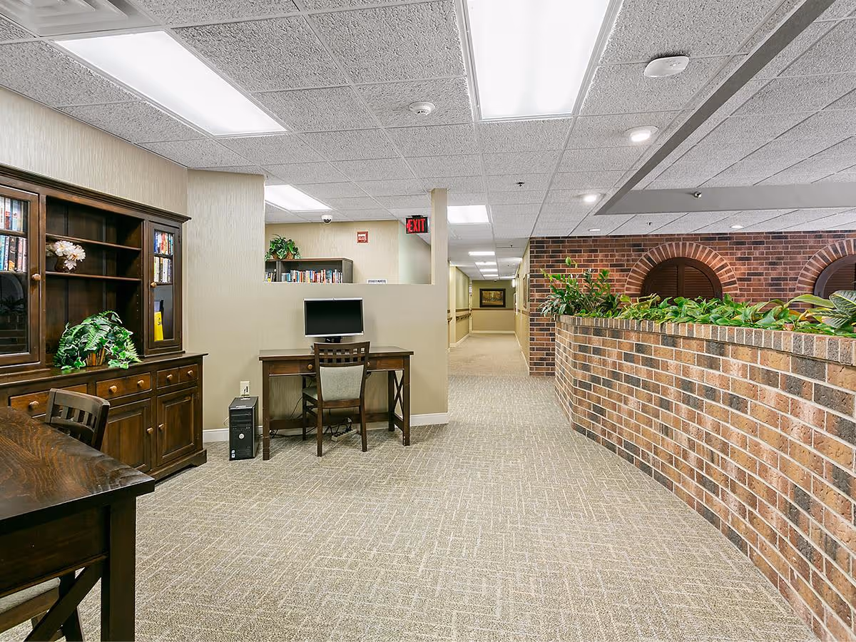 Interior view of a senior living facility hallway with beige carpet and walls. On the left side, there is a wooden desk with a computer and chair, and a wooden cabinet with shelves holding books and decorative plants. On the right side, there is a brick half-wall planter with green plants. The ceiling has recessed lighting and fluorescent panels.