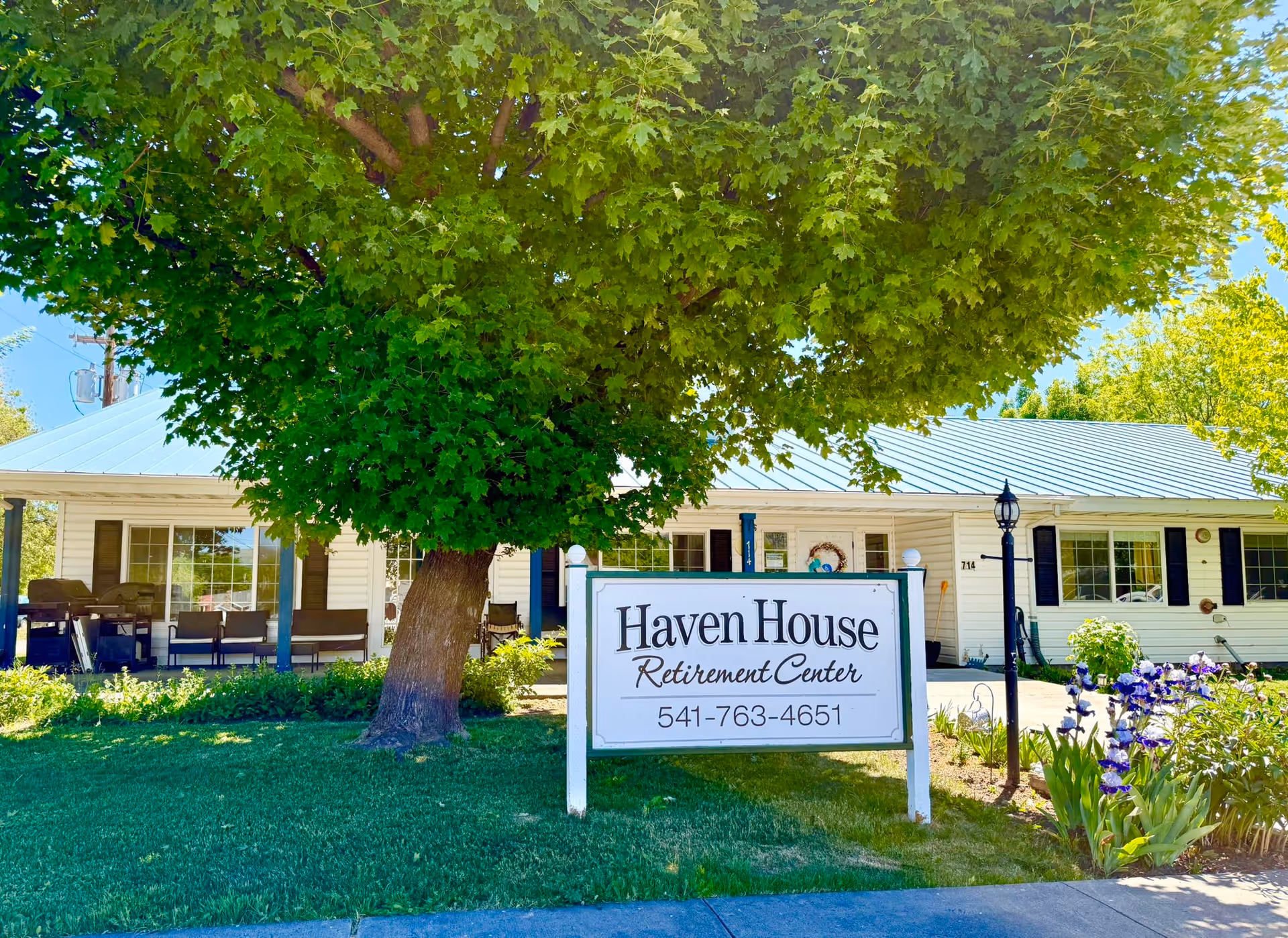 Front of a single-story retirement home with a large tree, lawn and a sign reading "Haven House Retirement Center" and a phone number.