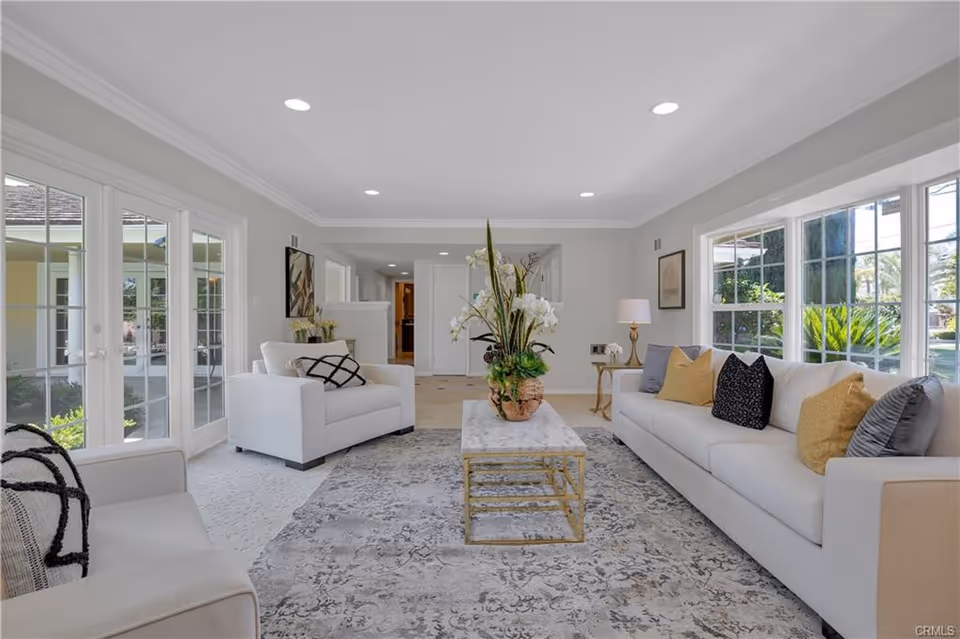 Bright modern living room with white sofas, a marble coffee table, large windows and glass doors, and decorative pillows and plants.