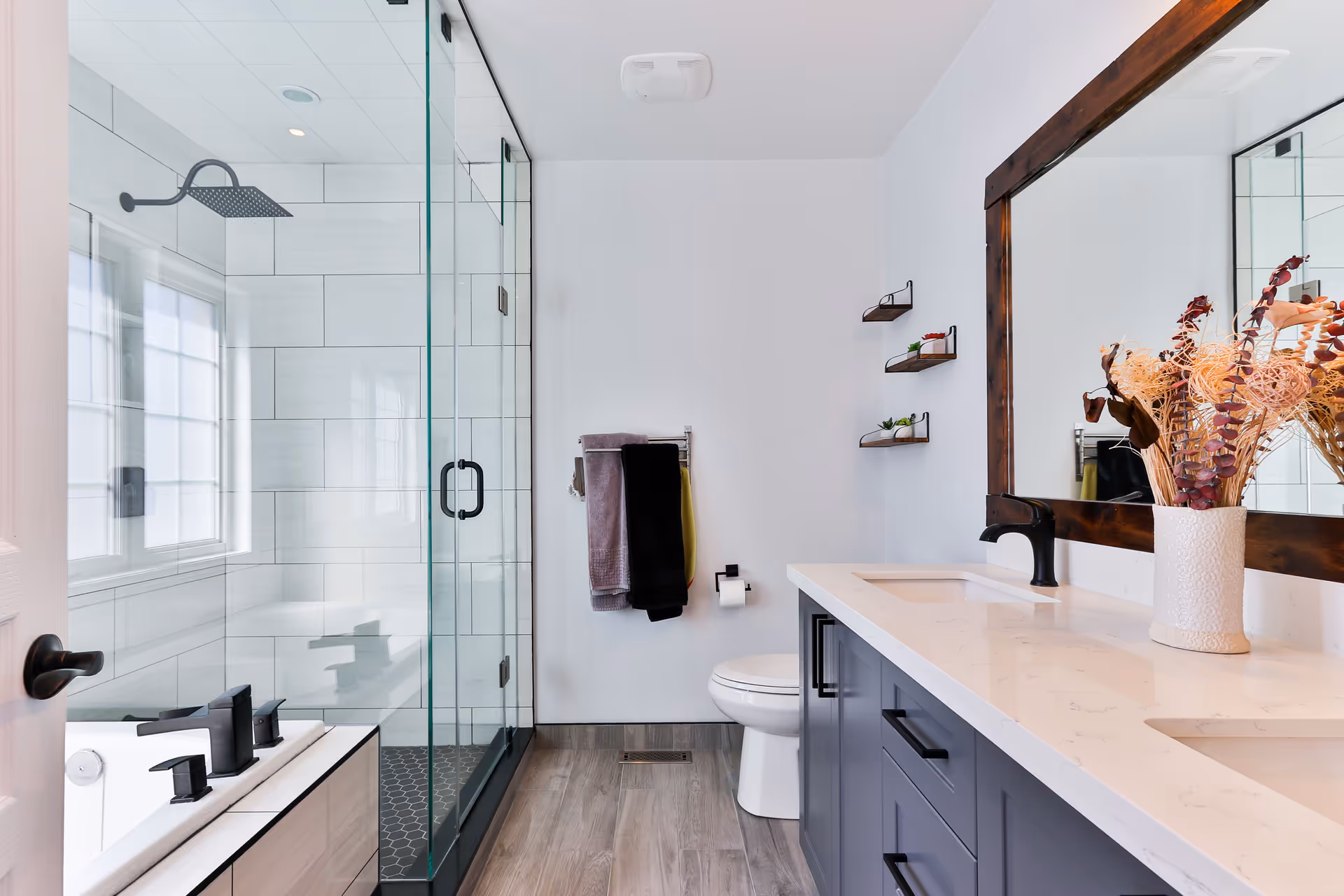 Modern bathroom with a glass-enclosed shower, a bathtub with black fixtures, a toilet, and a double sink vanity with a large wooden framed mirror and decorative vase with dried flowers. The walls are white, and the floor has wood-like tiles.