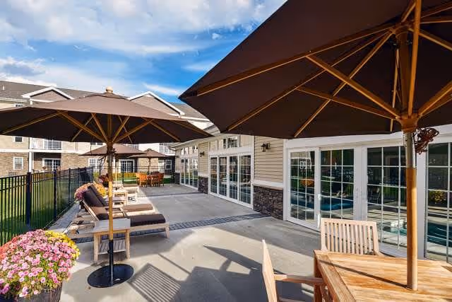 Outdoor patio area at Summit at Forts Ferry with lounge chairs, large umbrellas, wooden tables and chairs, and a flower pot with pink flowers. The patio is adjacent to a building with large windows and a fenced grassy area.