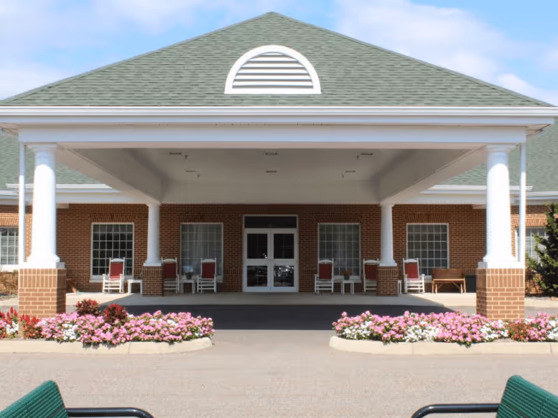 Entrance of a senior living facility with a covered driveway supported by white columns, red brick walls, several windows, and rocking chairs on the porch. Flower beds with pink and white flowers line the driveway.