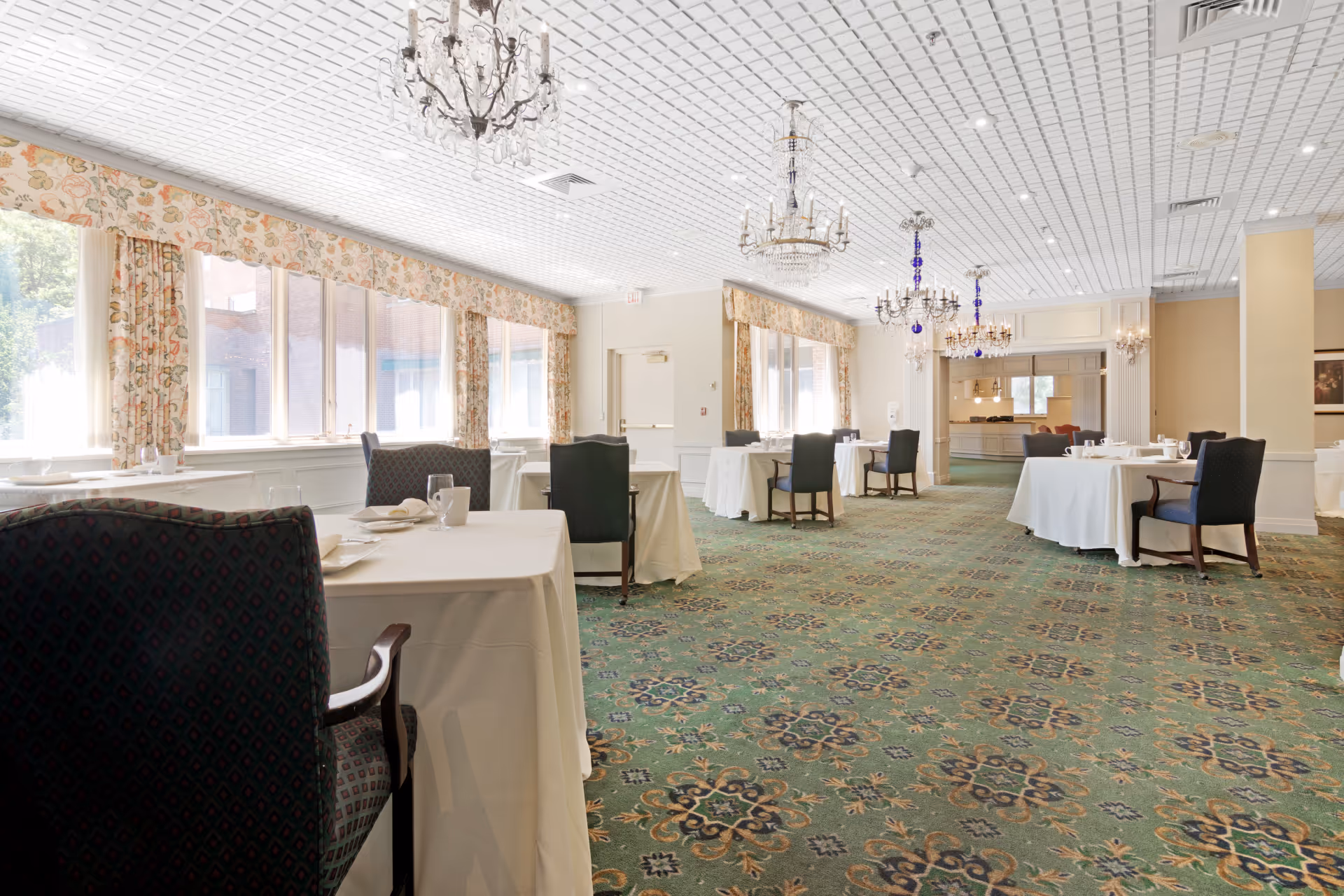 Spacious formal dining room with tables set with white tablecloths, upholstered chairs, chandeliers, and a patterned green carpet.