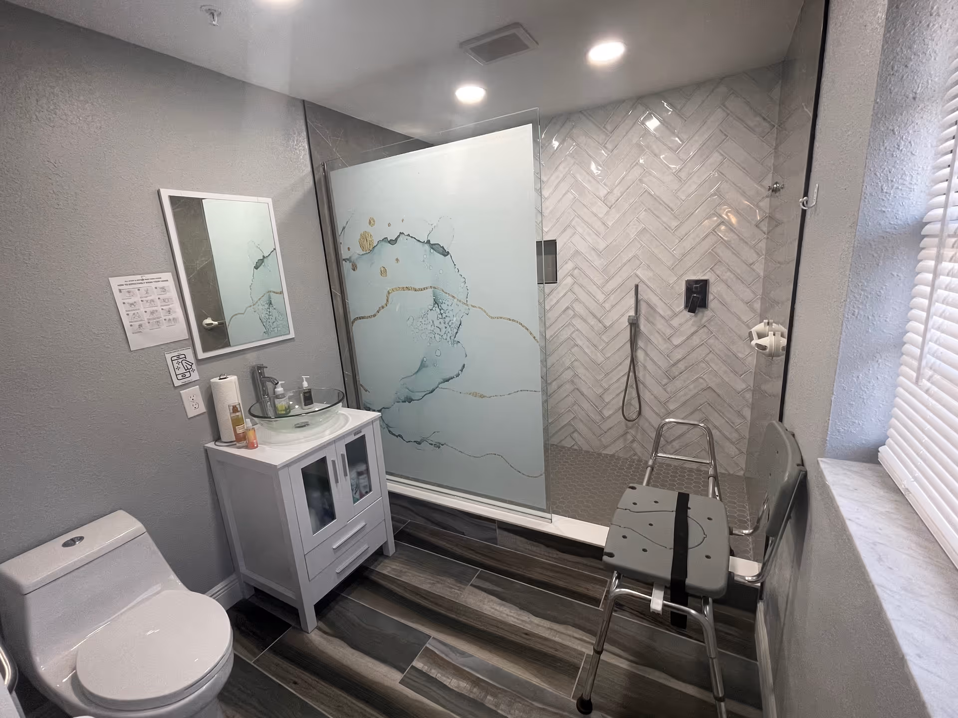 Accessible bathroom with glass shower, sink vanity, toilet, and a shower chair.