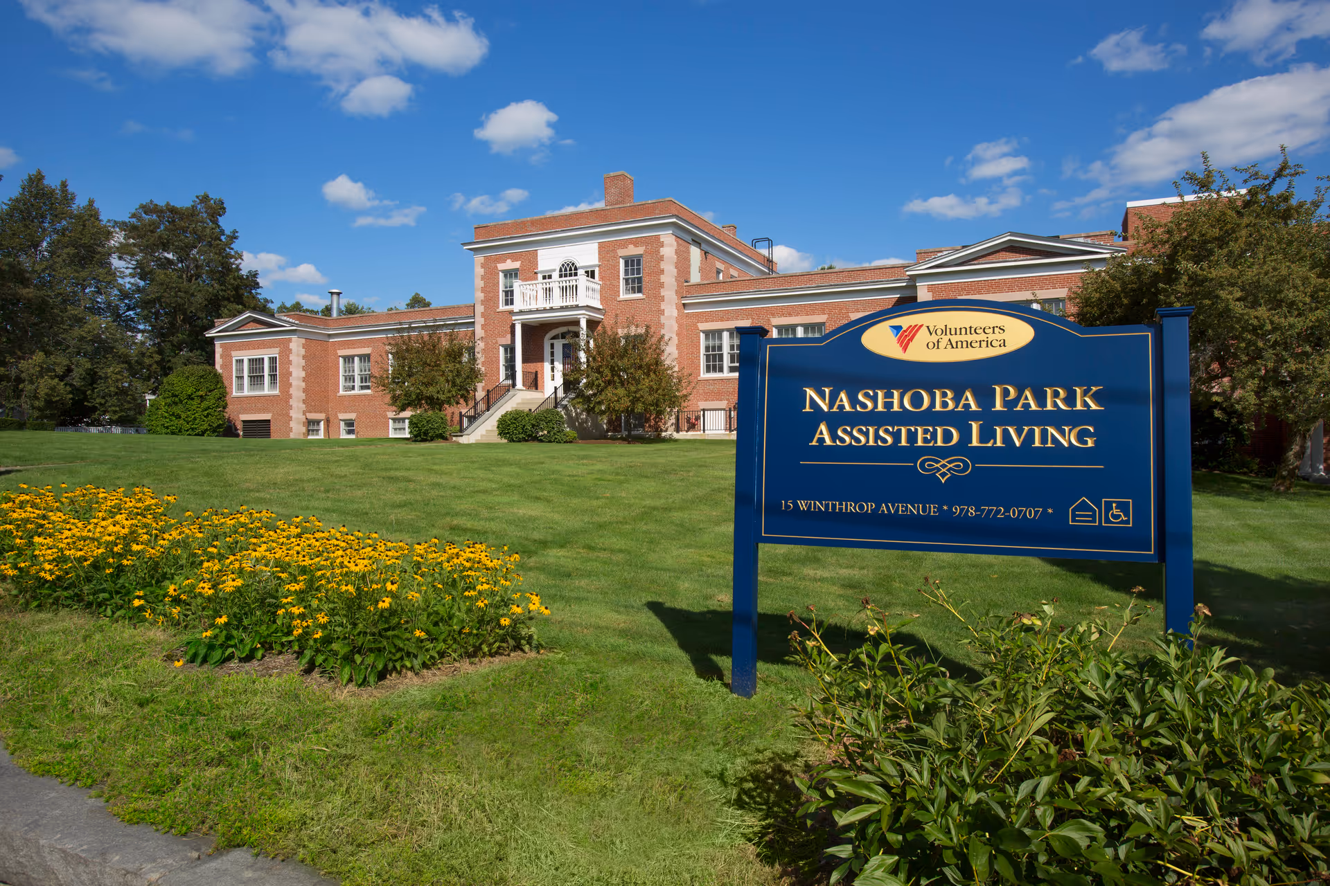Brick-assisted living building with a blue Nashoba Park sign on a green, sunny lawn.