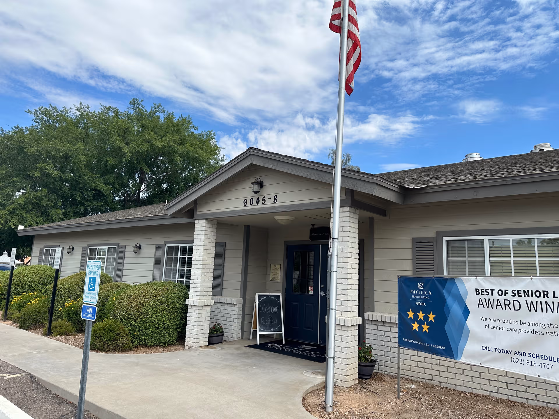 Single-story senior living facility entrance with flagpole, shrubs, and banners.