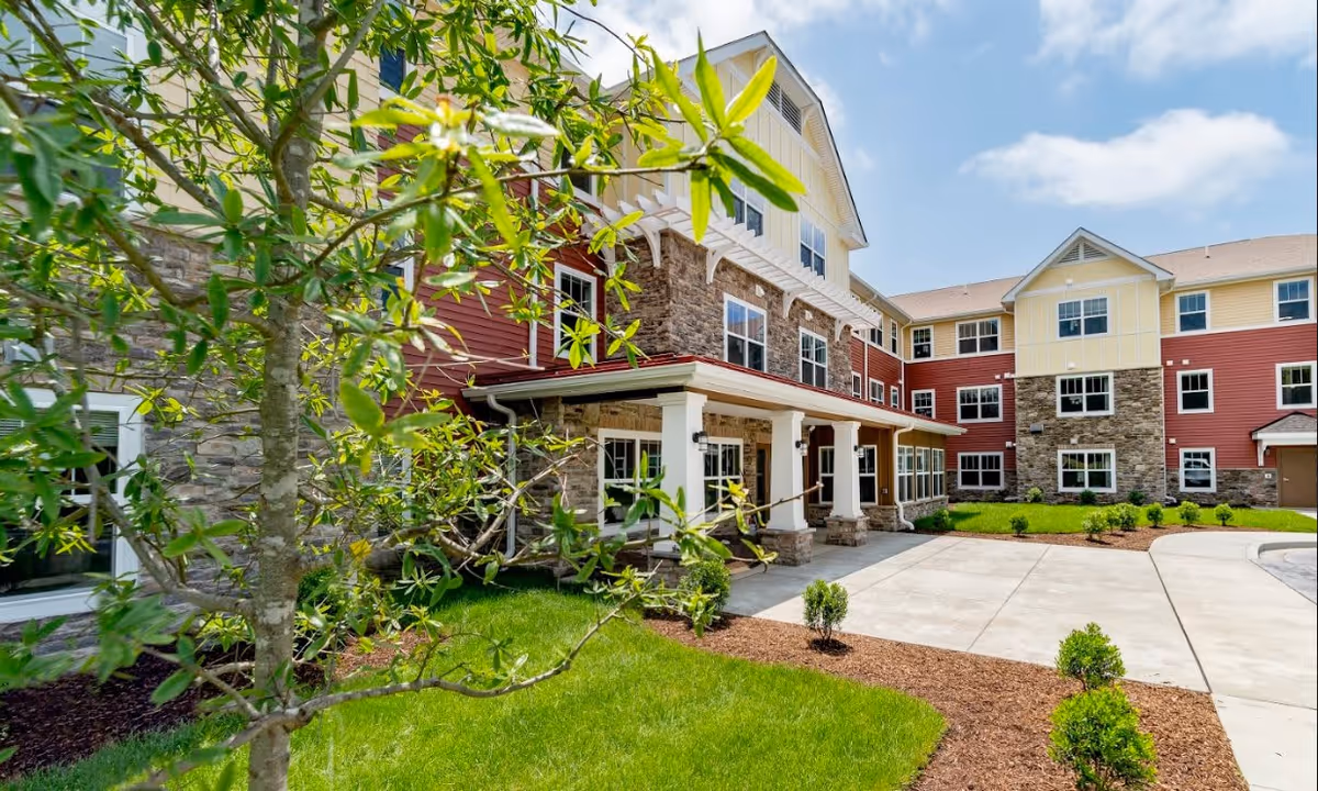 Front entrance and exterior of a multi-story senior living apartment building with red and yellow siding, stone accents, a driveway and landscaped greenery.