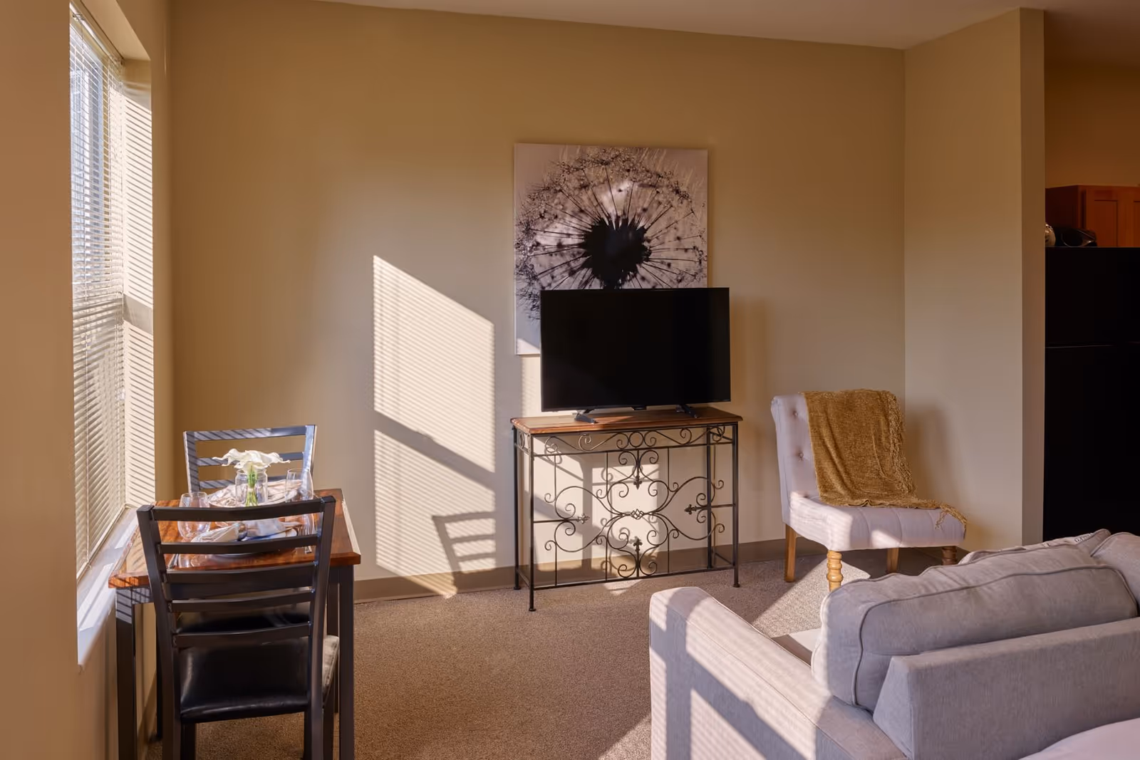 A cozy living room area with a small dining table set for two near a window with blinds. A flat-screen TV sits on a decorative metal and wood stand against a beige wall with a large dandelion artwork above it. There is a cushioned chair with a brown throw blanket and a light gray sofa partially visible in the foreground.