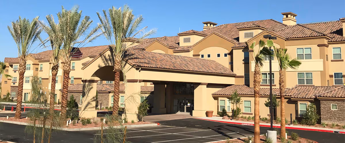 Exterior view of a large, multi-story retirement facility building with a covered entrance, surrounded by palm trees and landscaping under a clear blue sky.